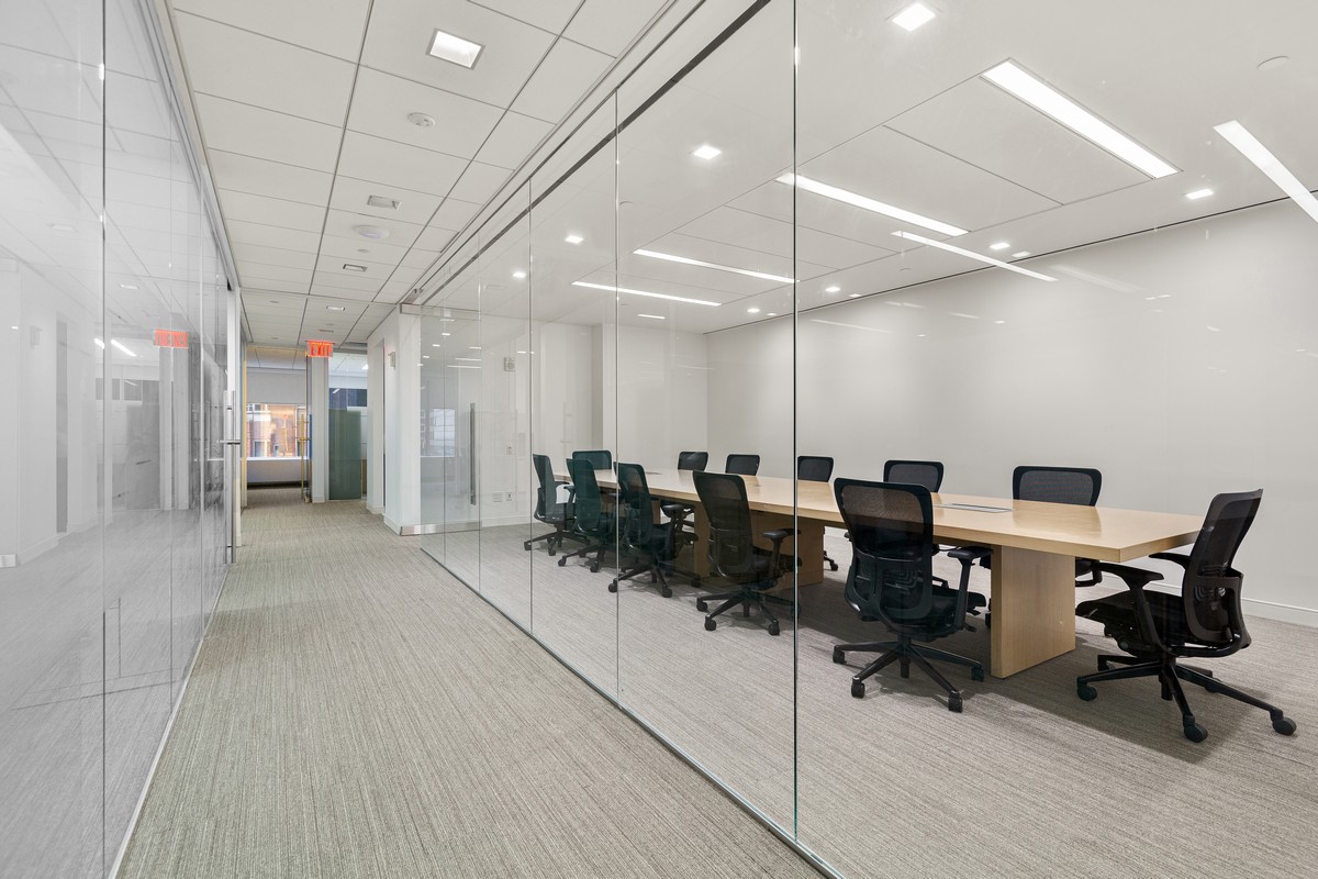 Glass-walled hallway past conference room with light wood table and mesh office chairs