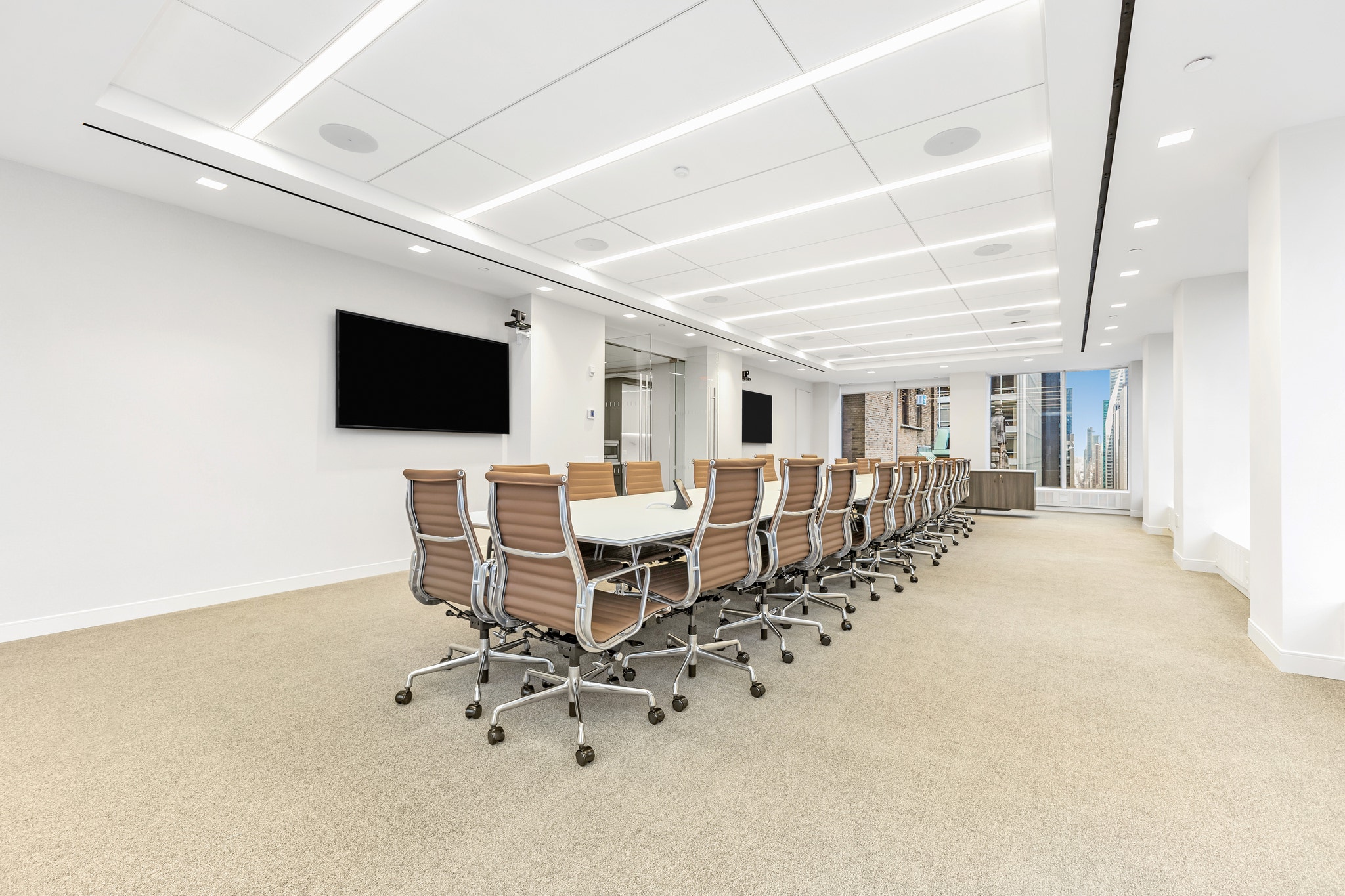 Long boardroom table with camel leather chairs, dual TV screens, and skyline window views