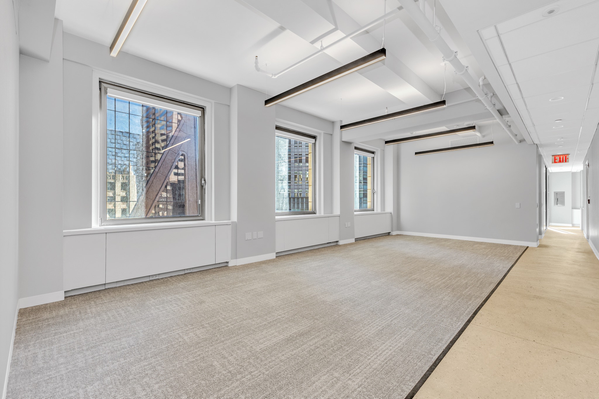 Empty corner office with gray carpet, three large windows, and angled ceiling light fixtures