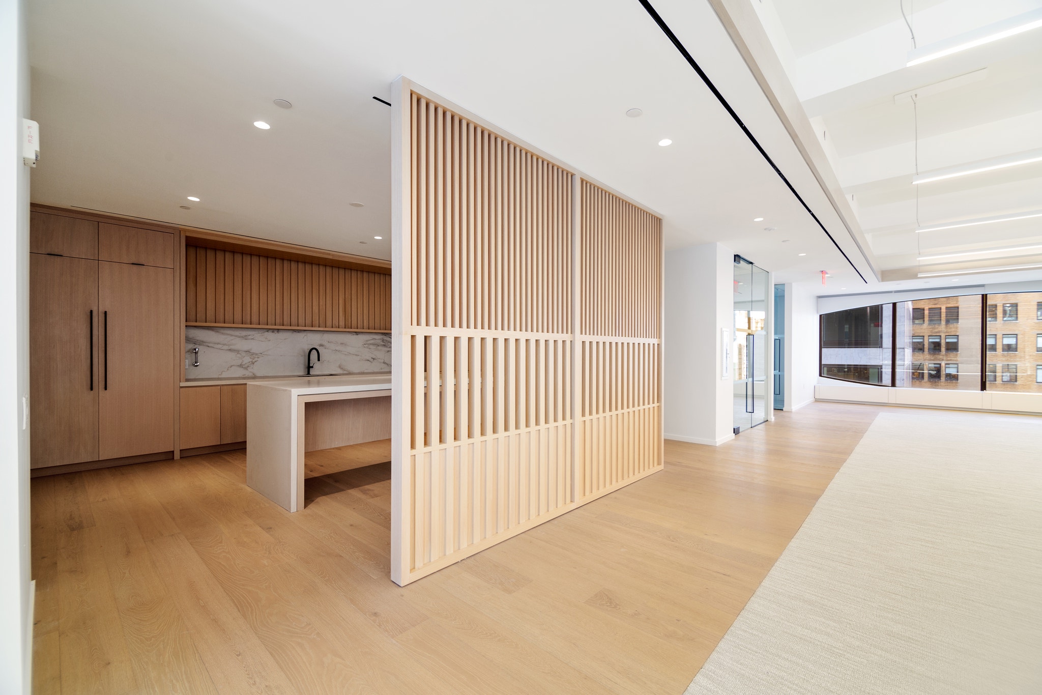 Open floor plan with slatted wood room divider beside kitchen and glass-walled office suite