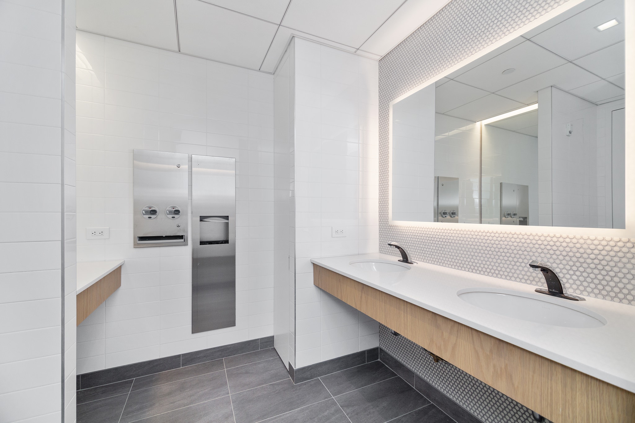 Modern restroom with dual sink vanity, penny tile backsplash, and backlit mirror wall