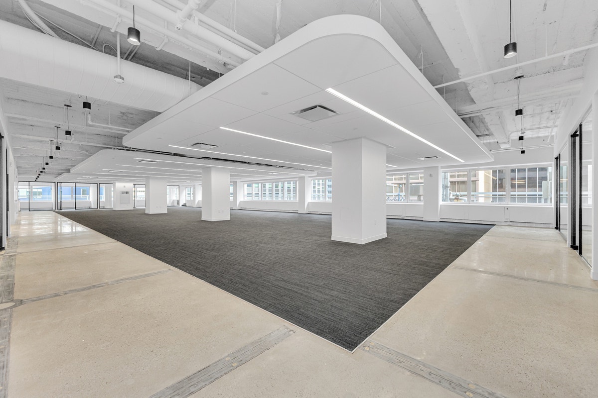 Open-plan office floor with dark gray carpet tile, exposed ductwork, and white structural columns