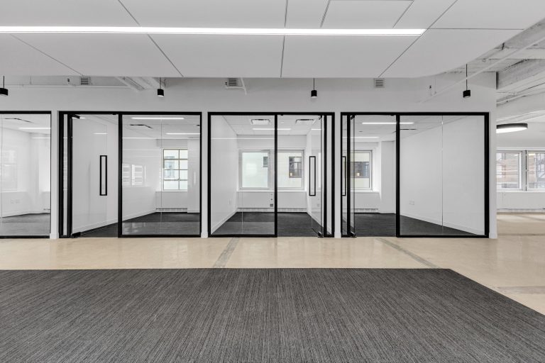 Row of empty glass-walled offices with black metal frames along a raw concrete and carpet floor