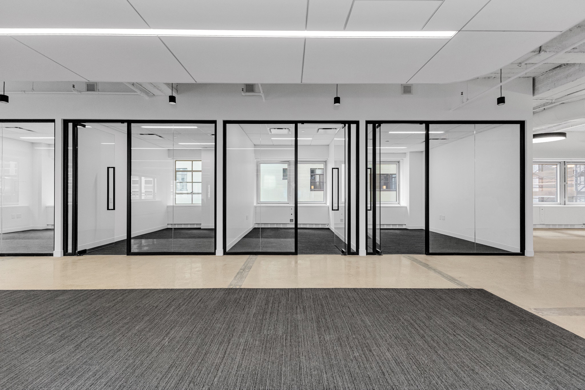 Row of empty glass-walled offices with black metal frames along a raw concrete and carpet floor