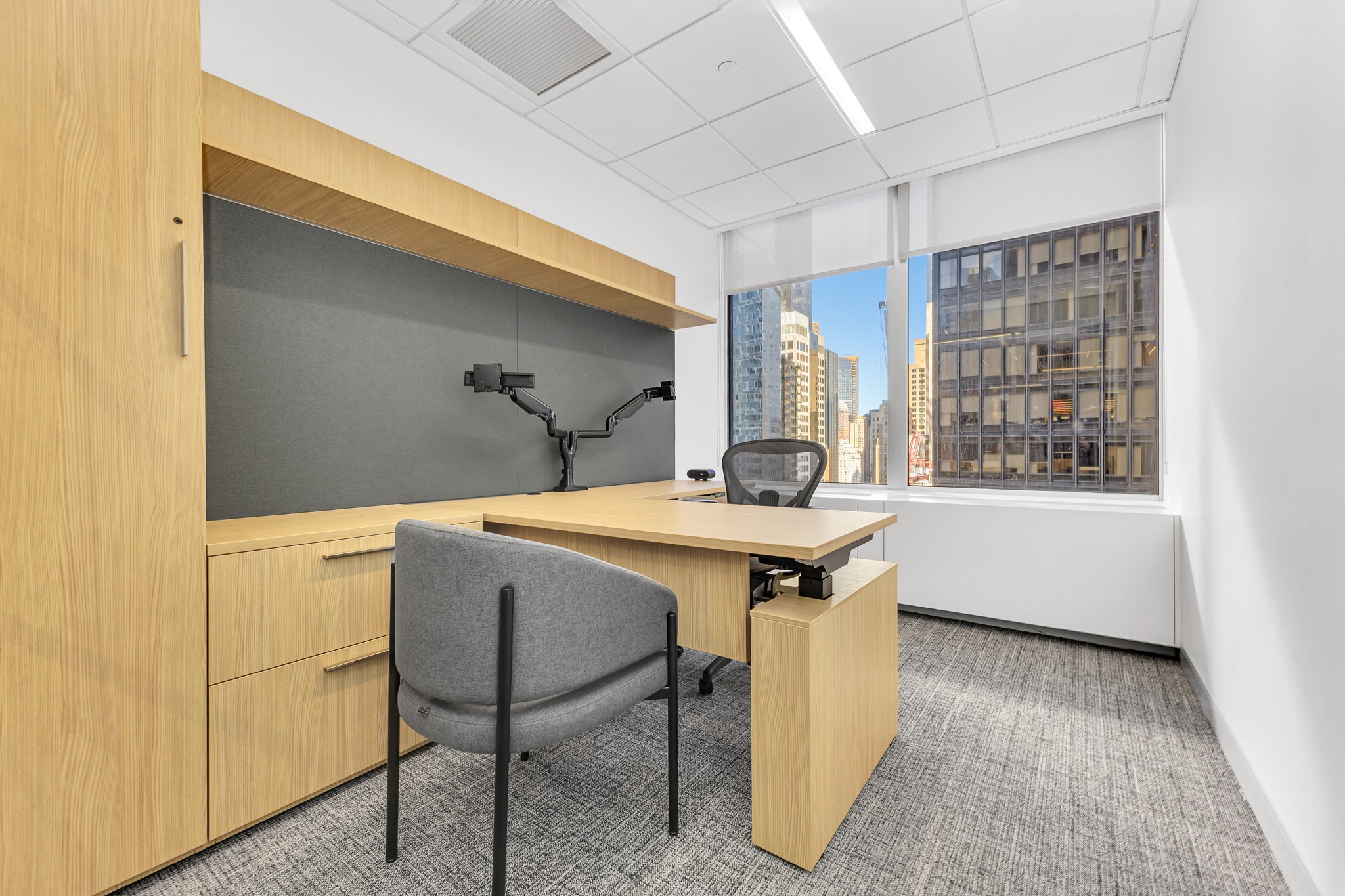 L-shaped wood desk office with dual monitor arm, gray task chair, and dense Manhattan skyscraper views