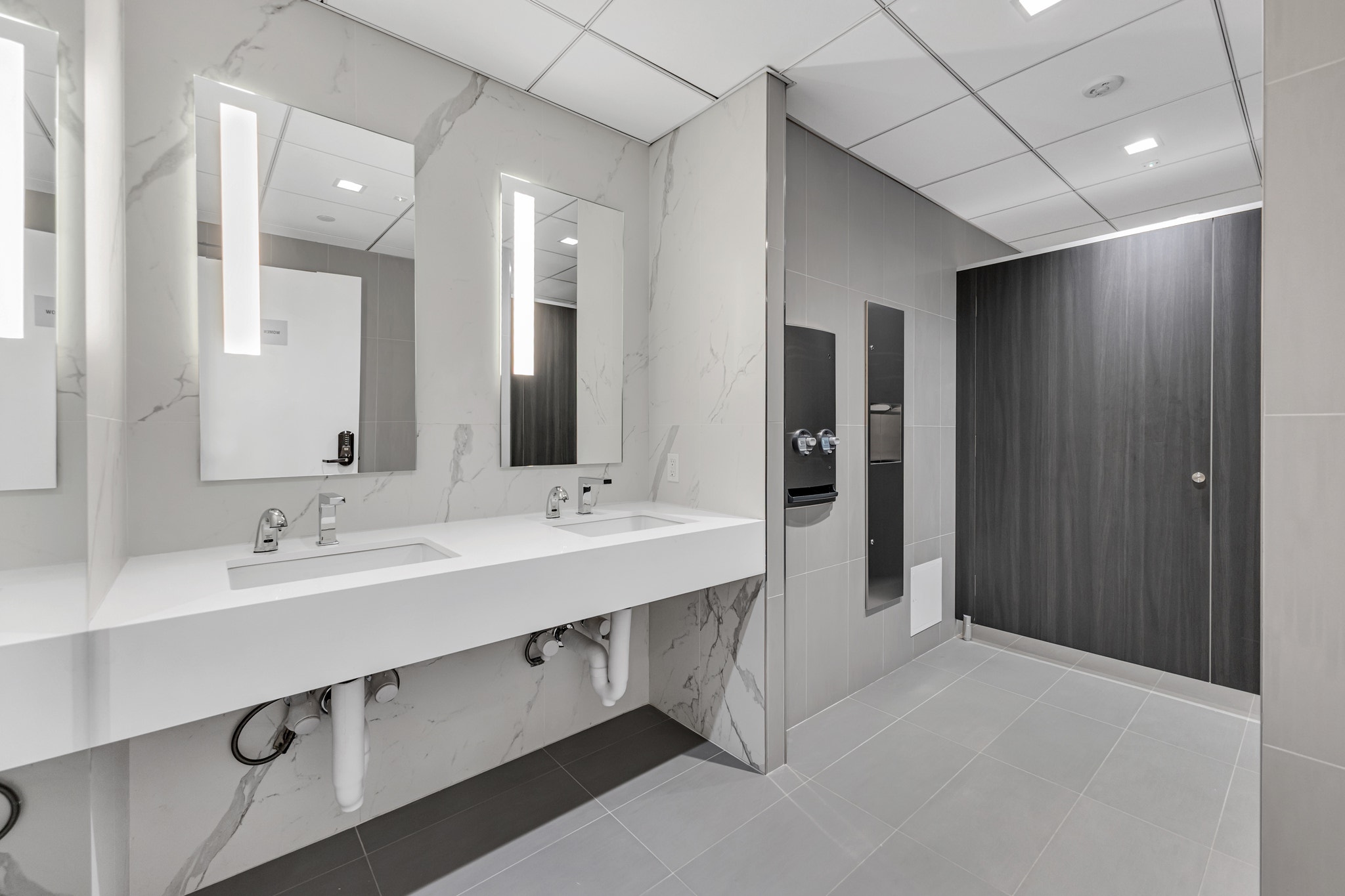 Office restroom with dual sink vanity, marble tile walls, backlit mirrors, and dark wood stall partition