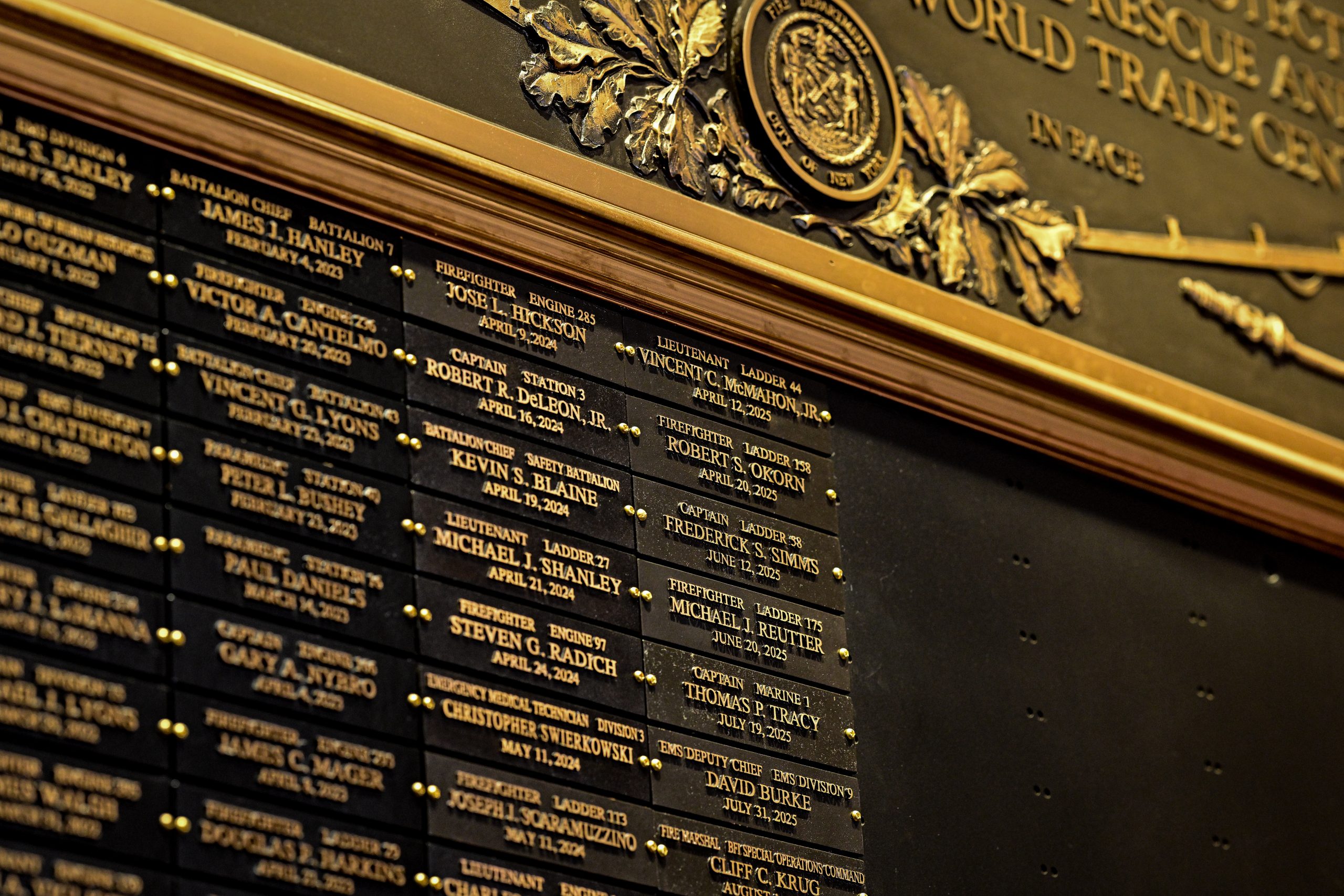 Close-up of a black memorial wall with gold-lettered plaques listing FDNY firefighters, officers, EMTs and paramedics by rank, unit, name and date of death, beneath an ornate gold FDNY seal and the words “World Trade Center” and “In Pace.”