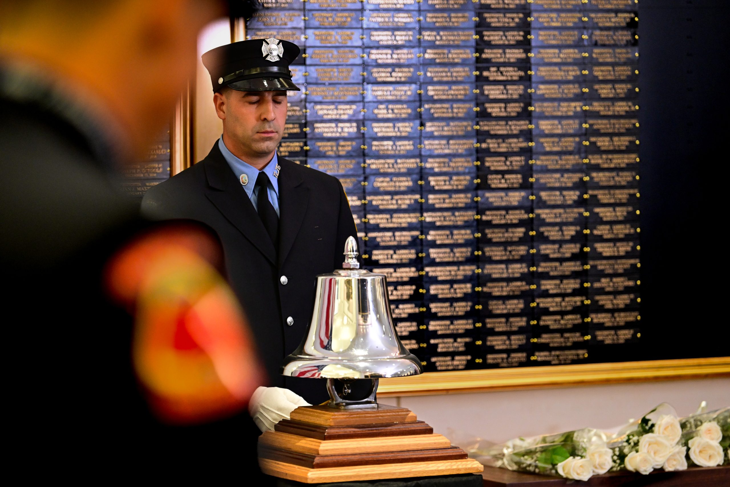 A uniformed FDNY firefighter in dress blues and white gloves stands with eyes closed beside a polished silver bell on a wooden base, in front of a wall of gold-lettered memorial plaques, with white roses laid nearby.