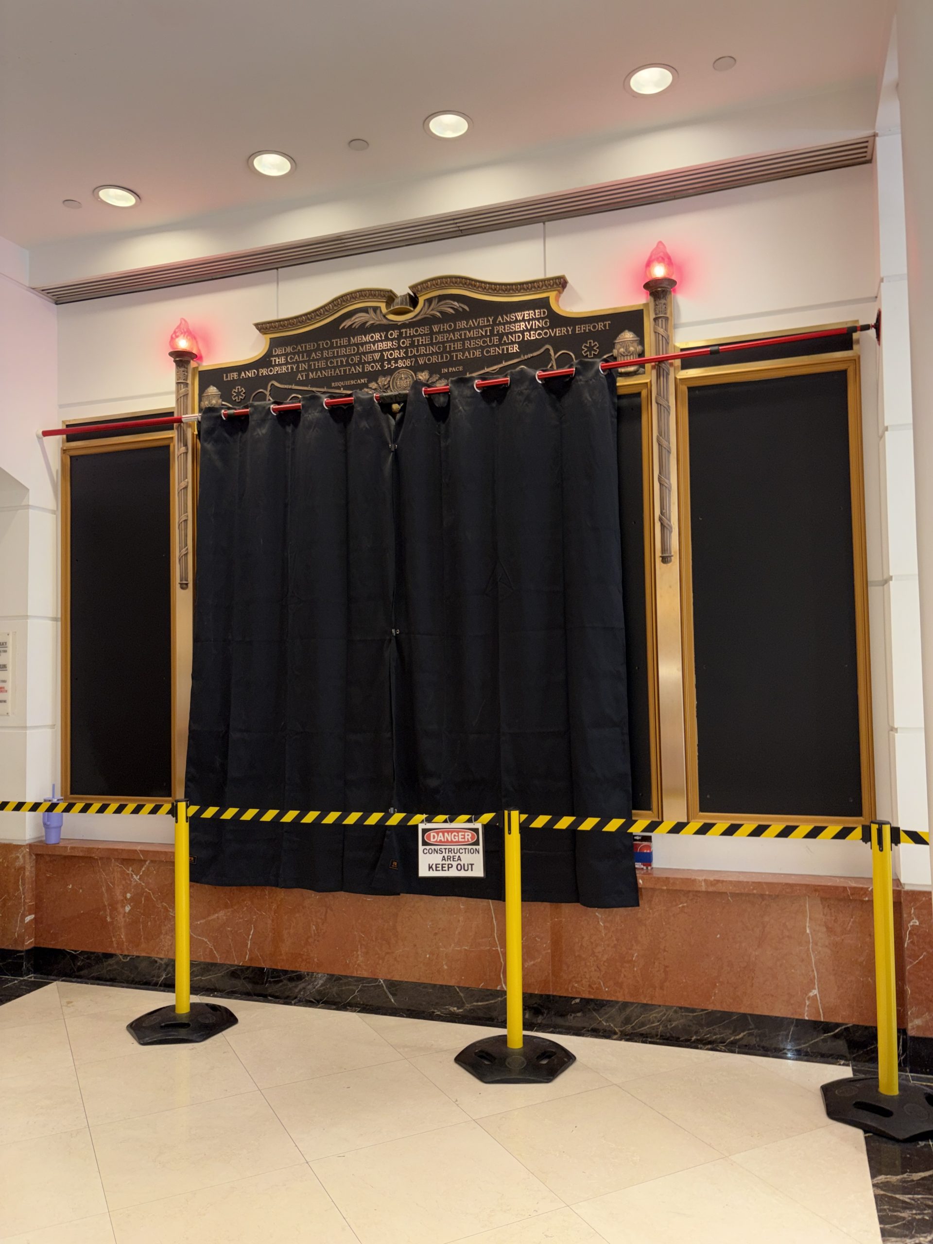An FDNY memorial wall partly covered by a black curtain hung from a red-and-white rod, with empty black panels on either side, two torch-shaped lamps glowing red, and yellow-and-black barrier tape with a “Danger: Construction Area, Keep Out” sign in front.